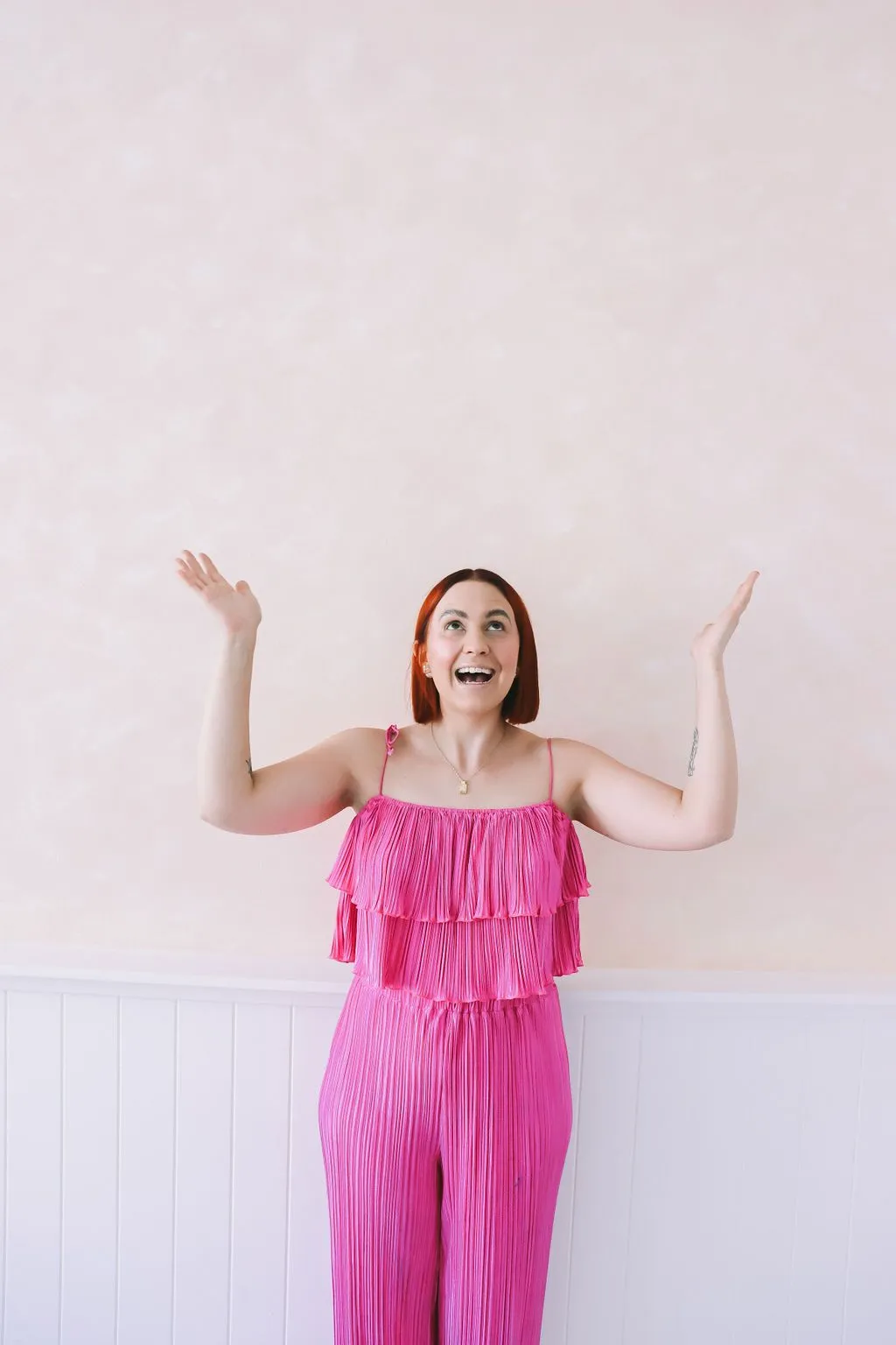 Katie in a pink jumpsuit celebrating with her arms raised